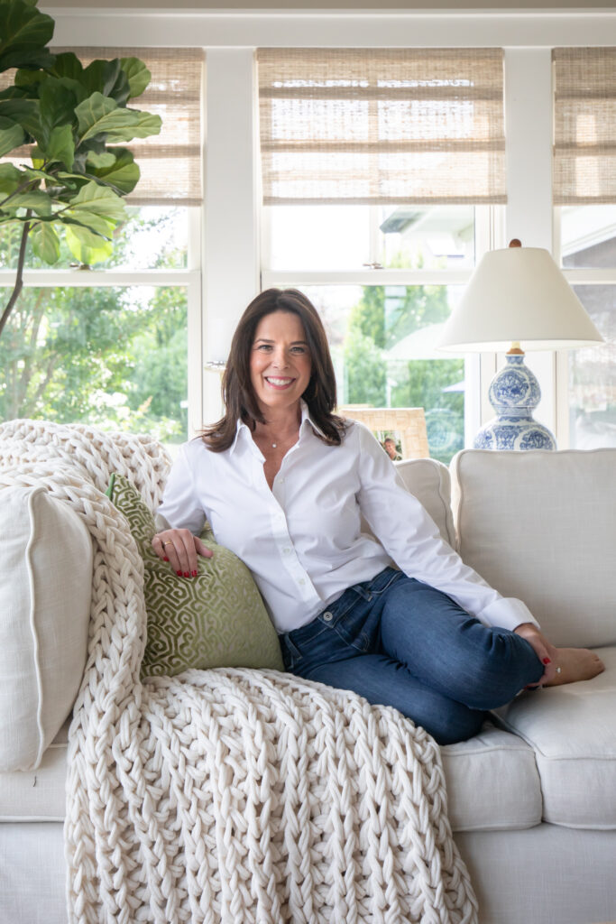 Jeanne Allen sits upon a sofa with green pillow in a bright sun room